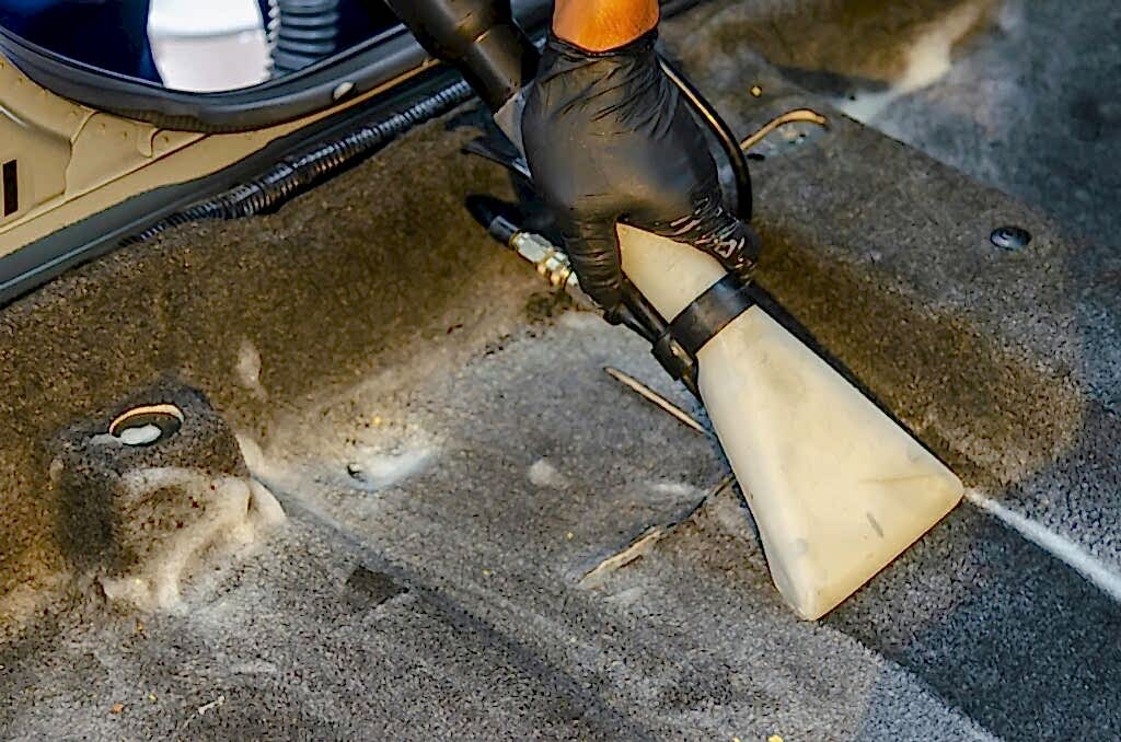 A hand in a glove using a vacuum to clean a car interior carpet, removing dirt effectively.